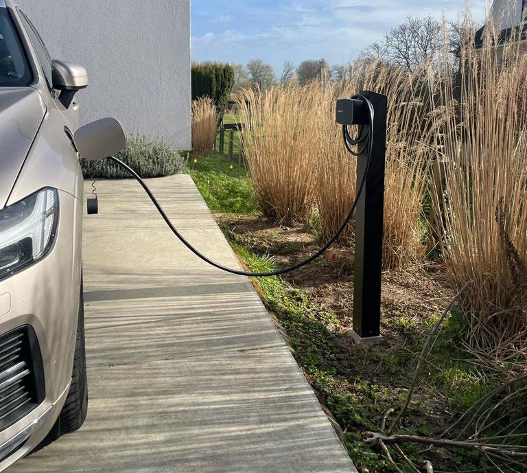 Elektrische wagen die thuis oplaadt aan een zwarte staande laadpaal naast de oprit.